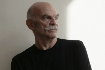 Medium-shot portrait of Martín Caparrós looking to the right, wearing a black shirt, sporting a gray mustache, against a light background.